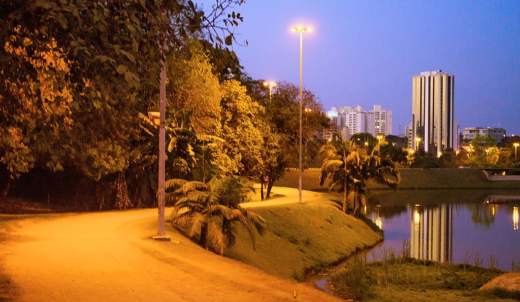 Uma estrada com curvas e um lago ao lado com prédios e céu azul ao fundo.